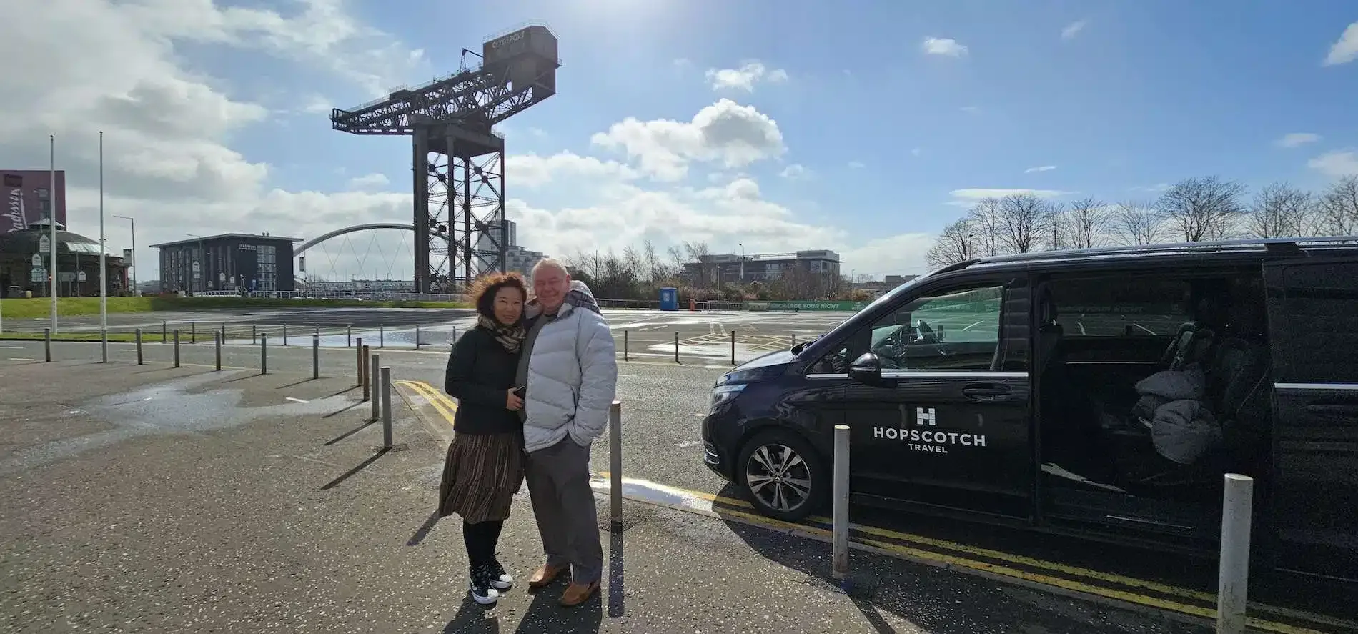 A couple standing near a black Hopscotch Travel van with Glasgow crane and bridge in the background.