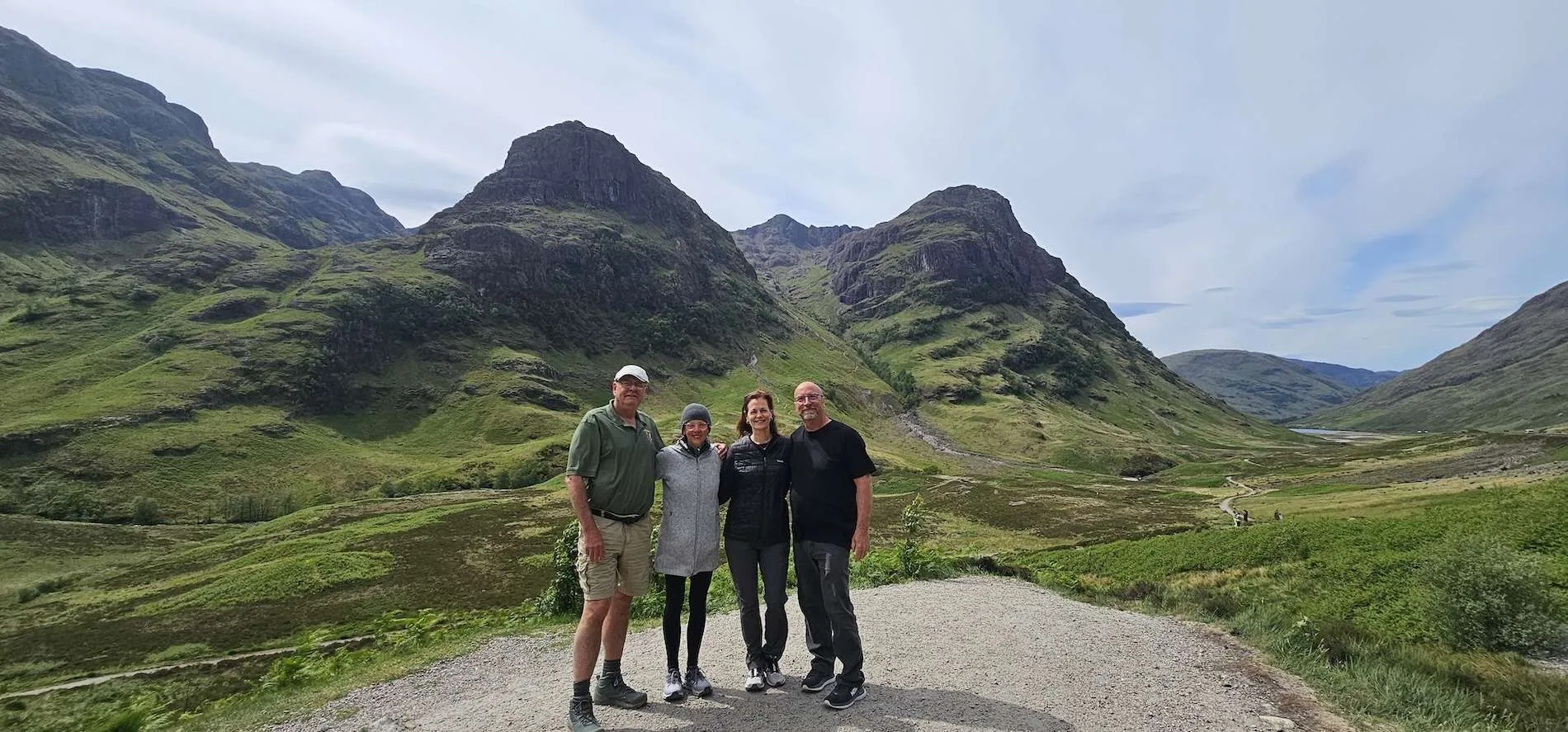 Four people standing on a gravel path with green mountains and cloudy sky behind them.