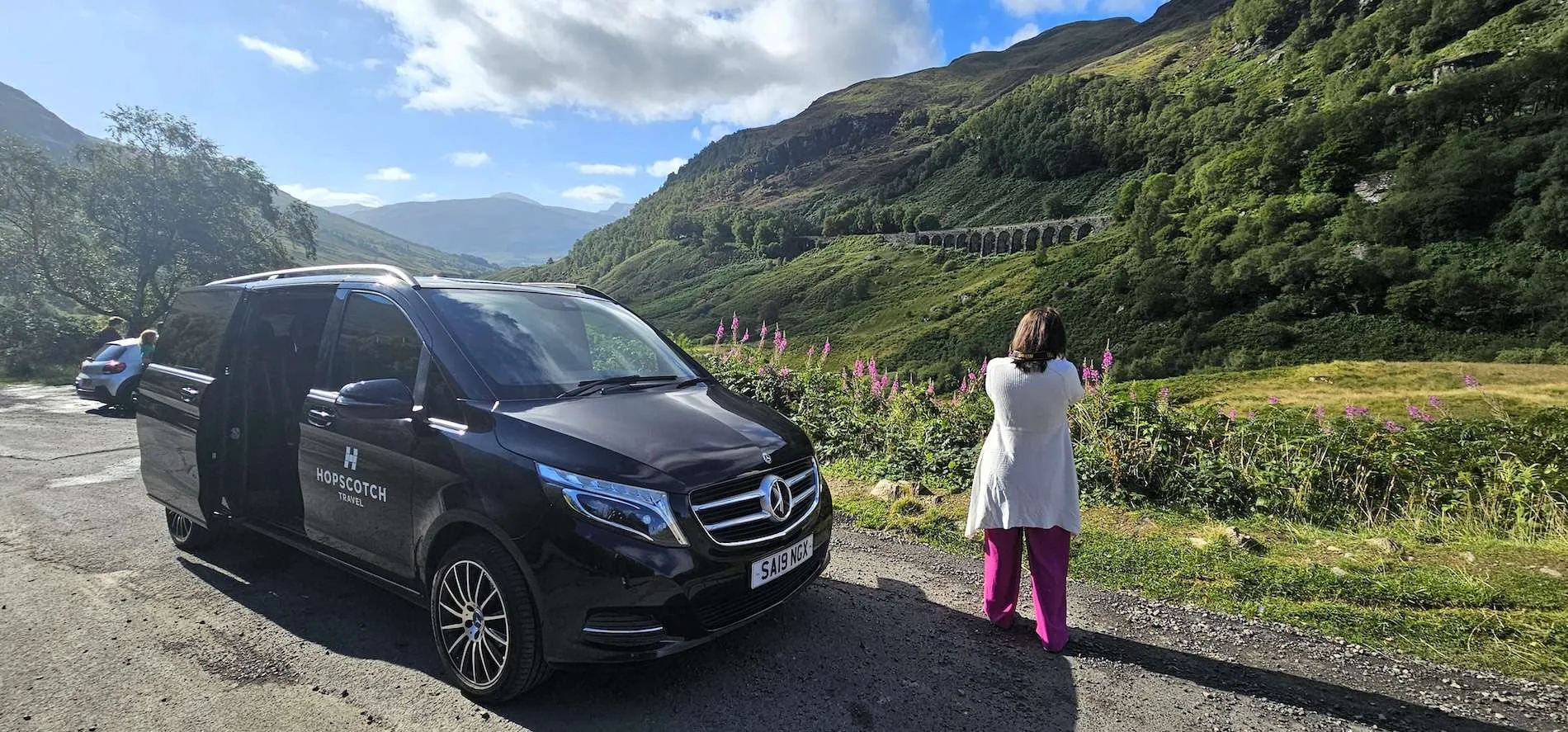 Black Hopscotch Travel van parked near a woman photographing a green valley and stone railway viaduct.