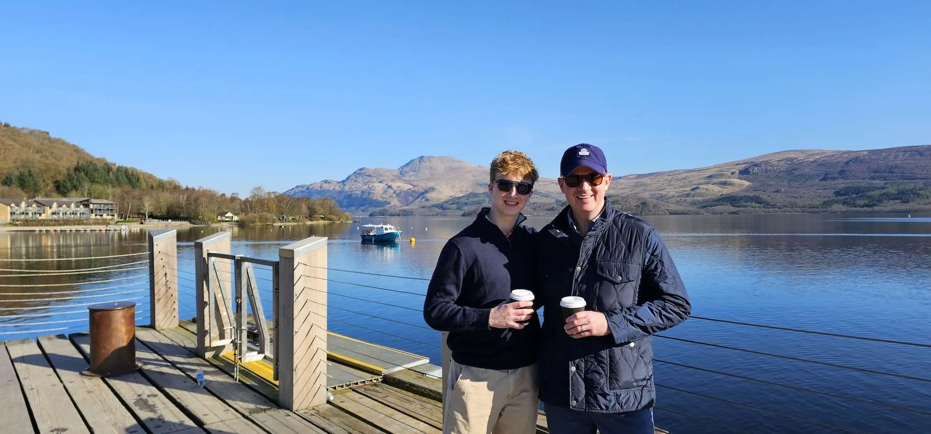 Two men in jackets and sunglasses holding coffee cups on a dock by a lake with hills and a boat in the background.