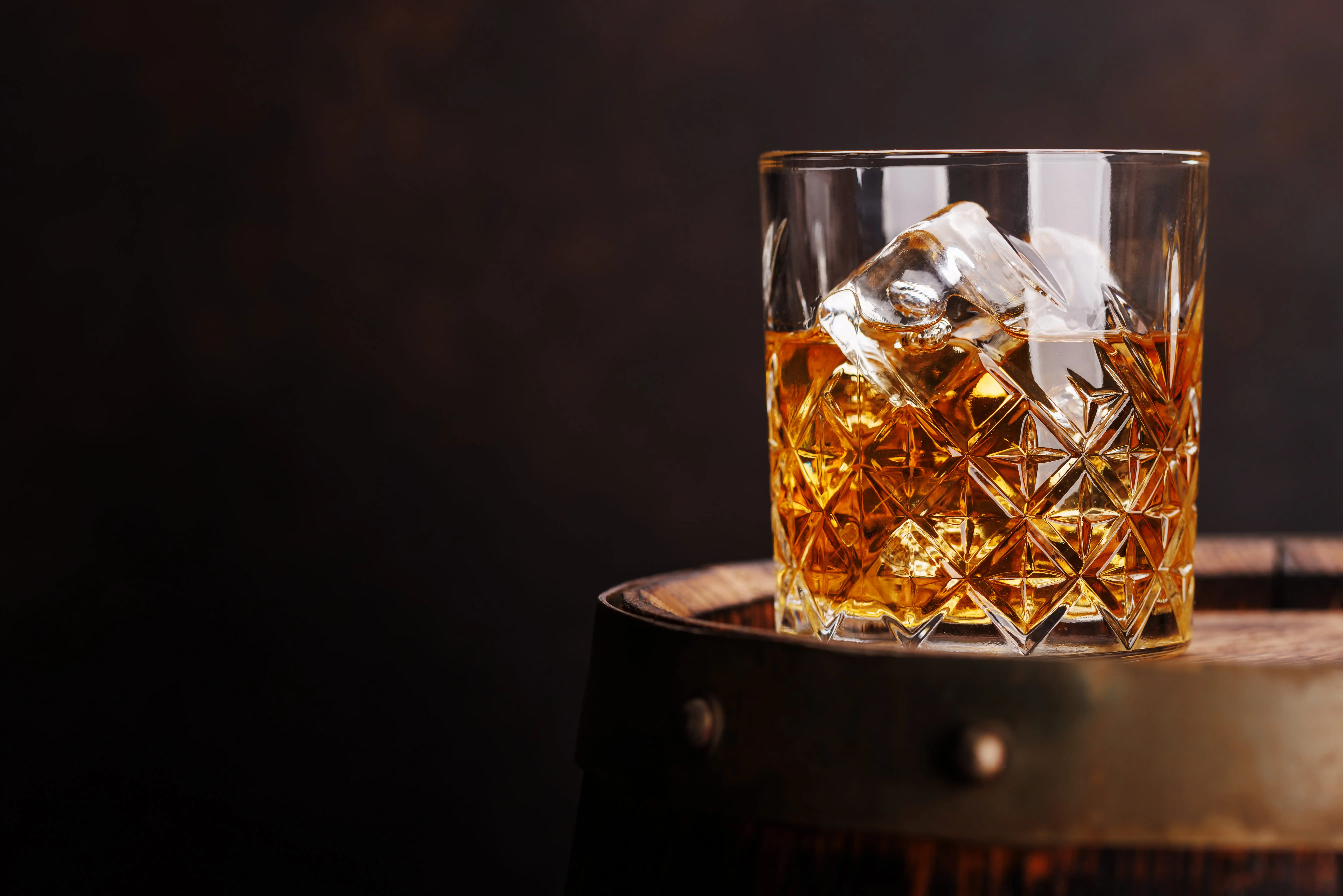 Glass of whiskey with ice cubes on top of a wooden barrel against a dark background.