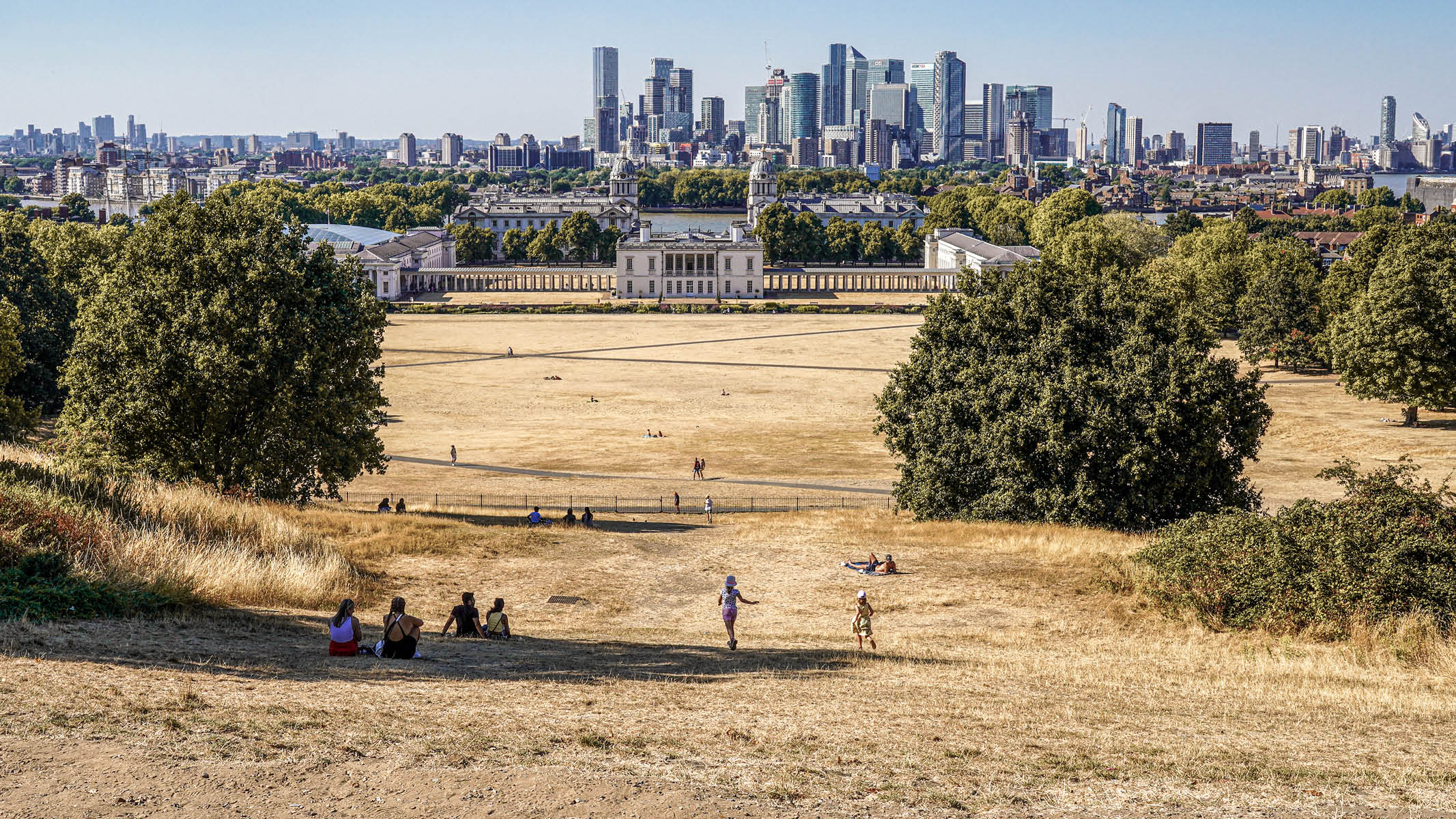 View of a dry grassy park with people sitting and walking, historic buildings, and a city skyline.