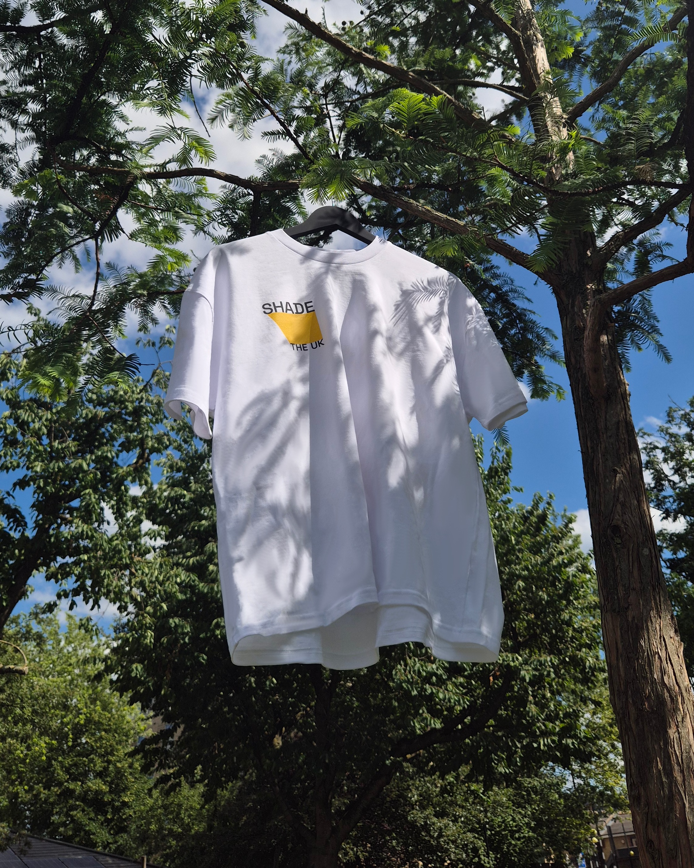 White T-shirt with yellow shape and 'SHADE THE UK' text hanging on a hanger from a tree branch.