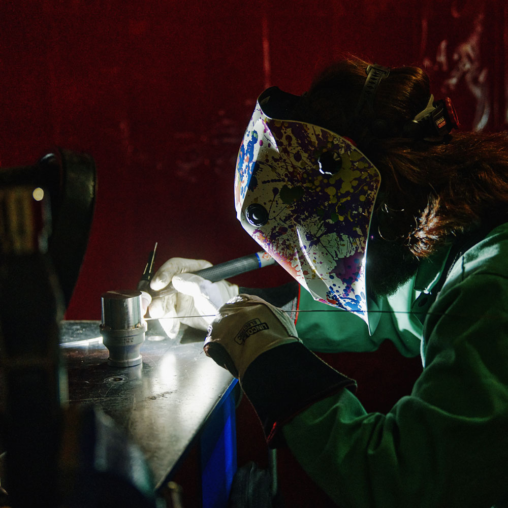 Person wearing a colorful welding helmet and gloves welding metal on a workbench with sparks.