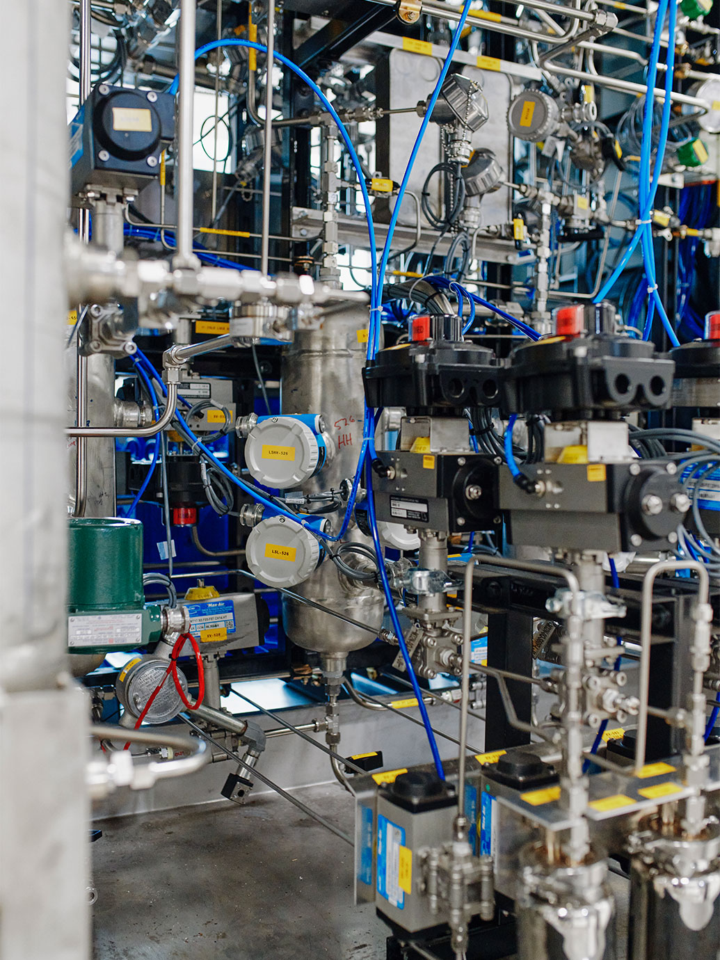 Complex industrial machinery with valves, gauges, metal pipes, and blue wiring in a factory setting.