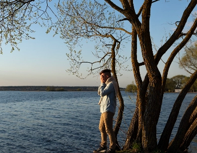 Man standing near a tree on a riverbank, talking on a phone at sunset.