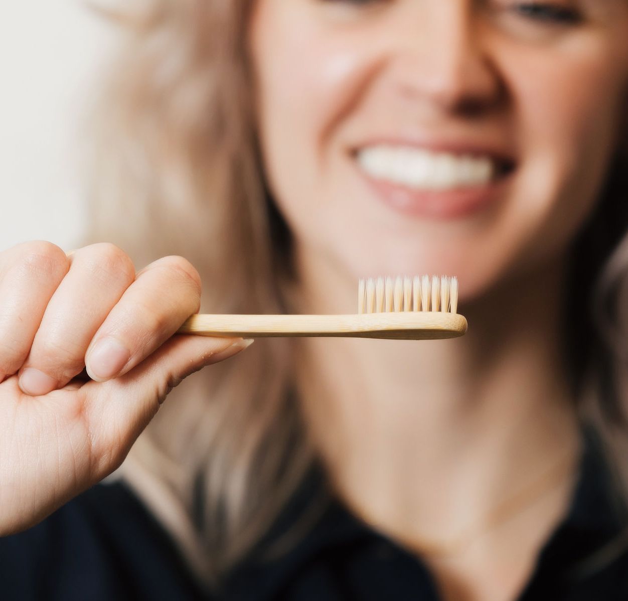 Woman smiling while holding a light wooden toothbrush with soft bristles close to the camera.