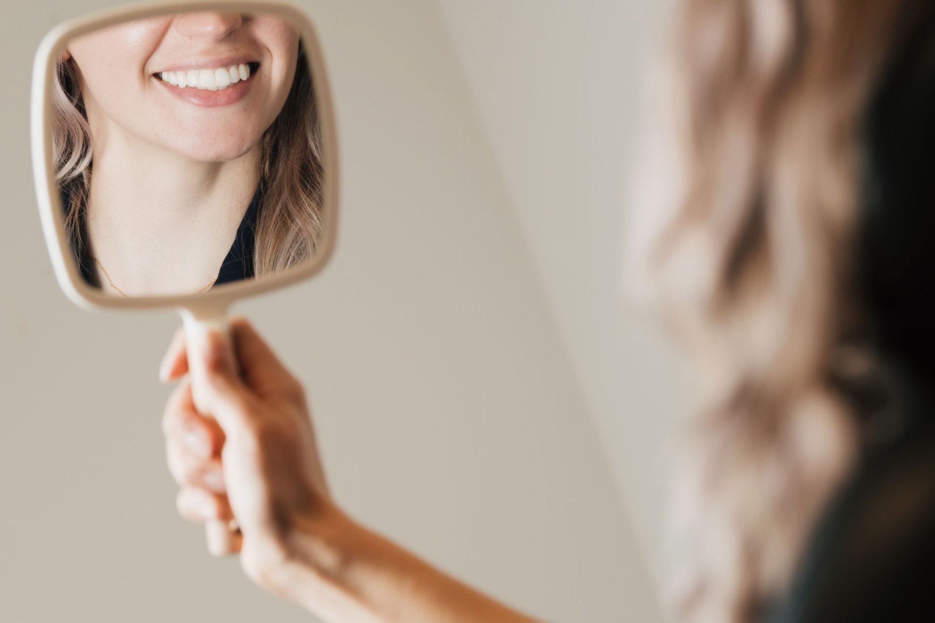 A woman holding a handheld mirror reflecting her smiling mouth and chin.