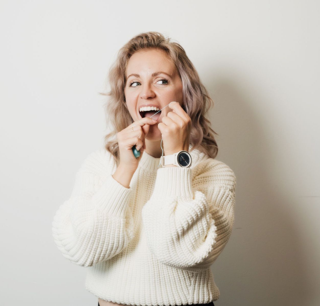 Woman in white sweater flossing her teeth with a green floss holder against a plain wall.