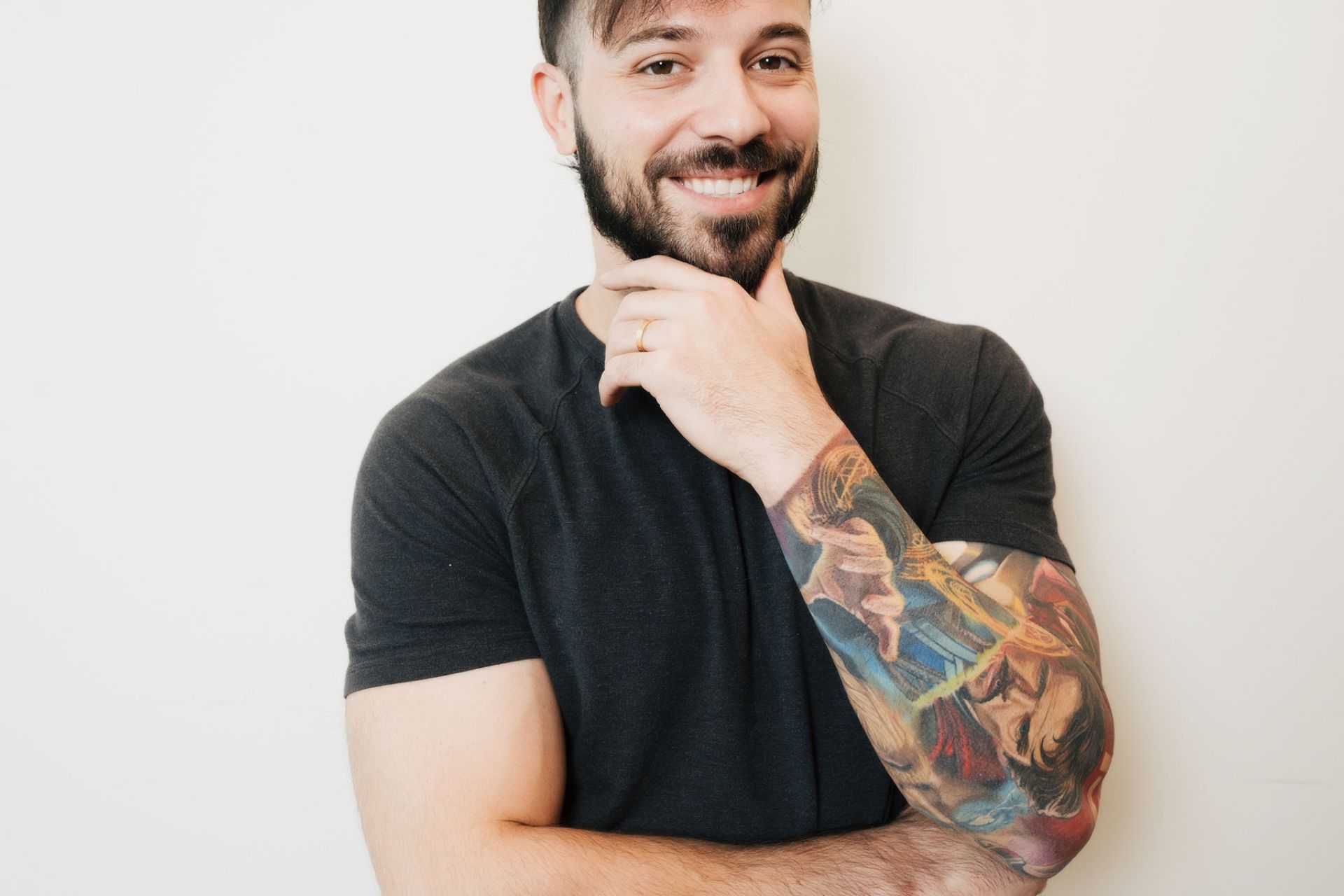 Smiling bearded man with a colorful superhero tattoo on his left forearm wearing a black short-sleeve shirt.