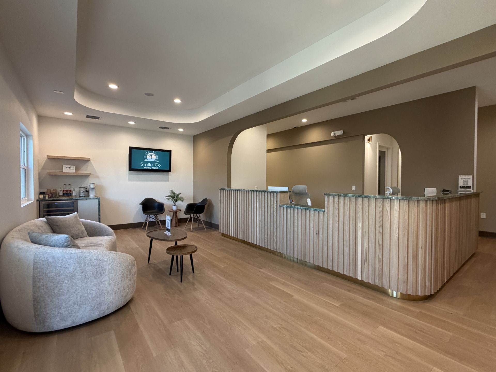 Modern waiting room with curved sofa, two chairs, wooden reception desk, and TV on white wall.