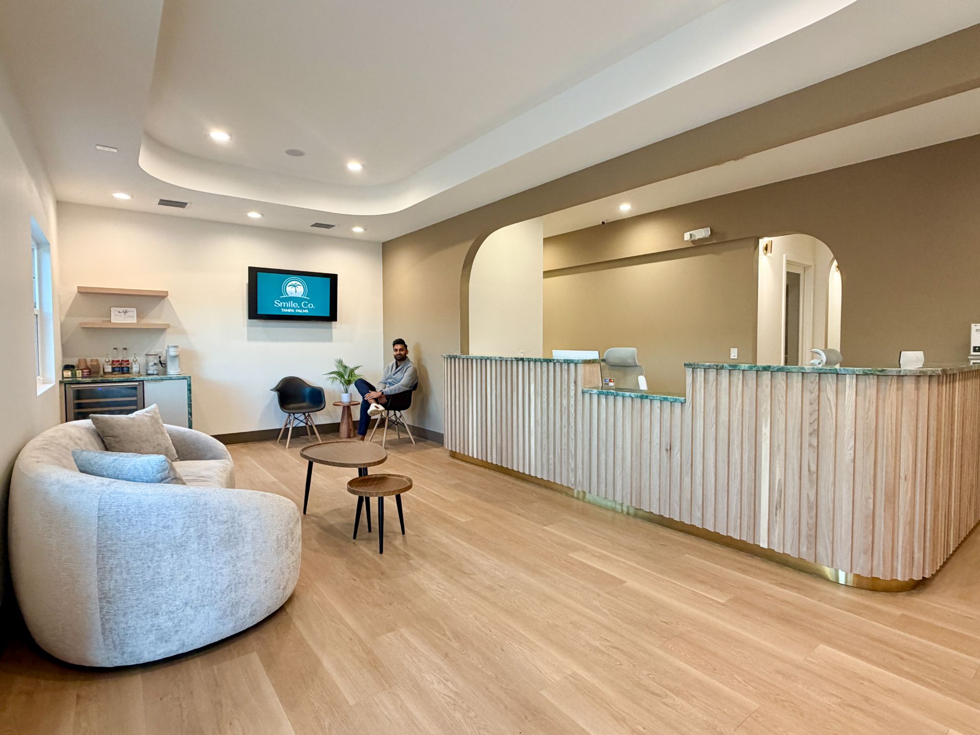 Modern reception area with wood desk, beige walls, a couch, and a man sitting on a black chair.