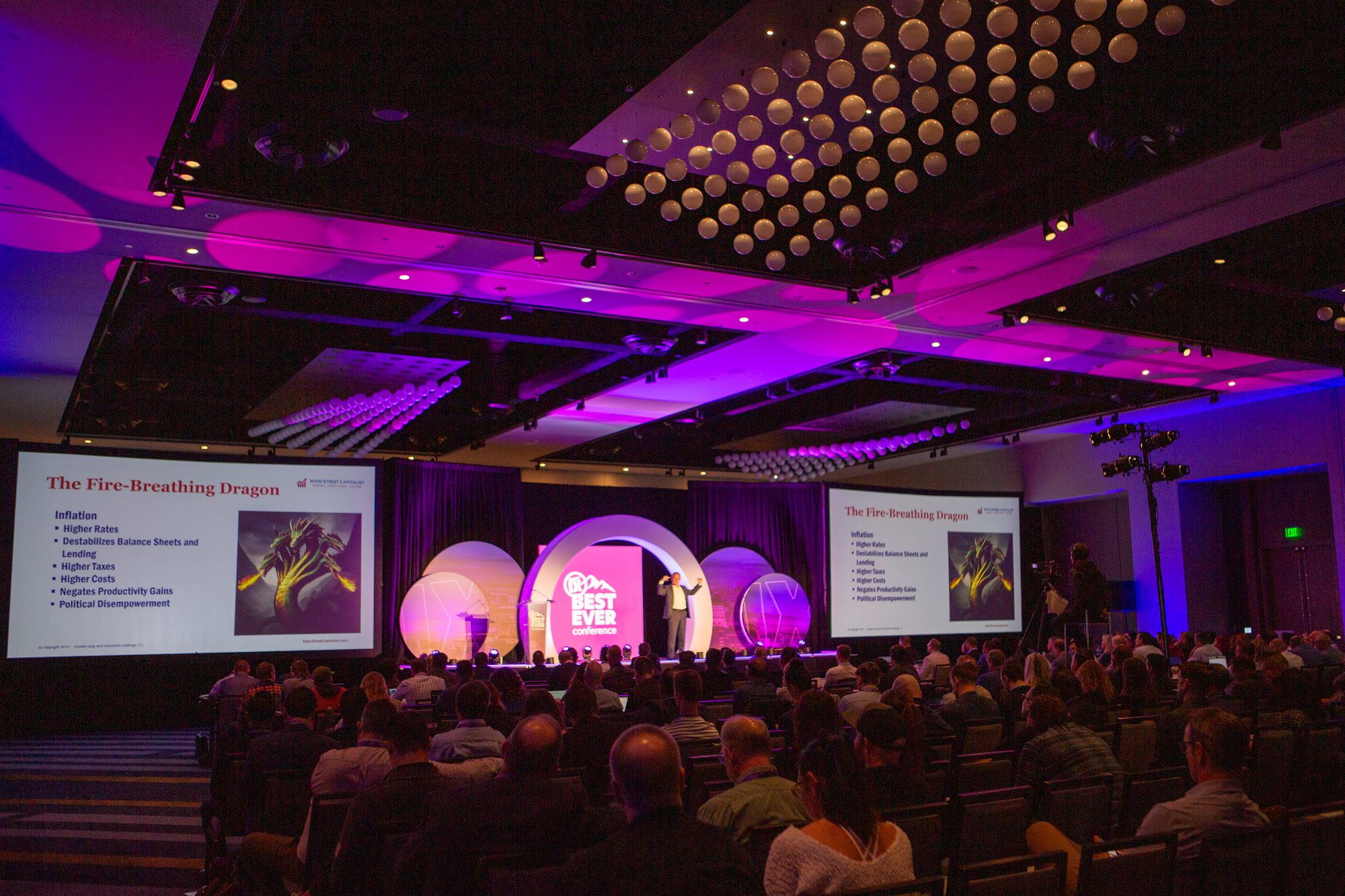 Keynote session in a full conference ballroom, stage lit in magenta and blue
