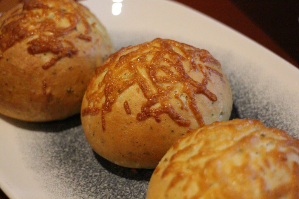 A cheese topped, round herb biscuit sits surrounded by others out of focus on a white and gray speckled plate