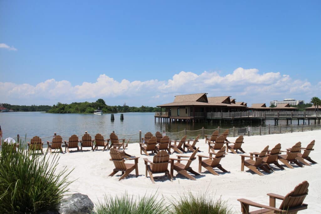 Polynesian resort beach chairs facing Cinderella Castle and the Seven Seas Lagoon. The sand is white, the water is calm and reflective and the sky is very blue.