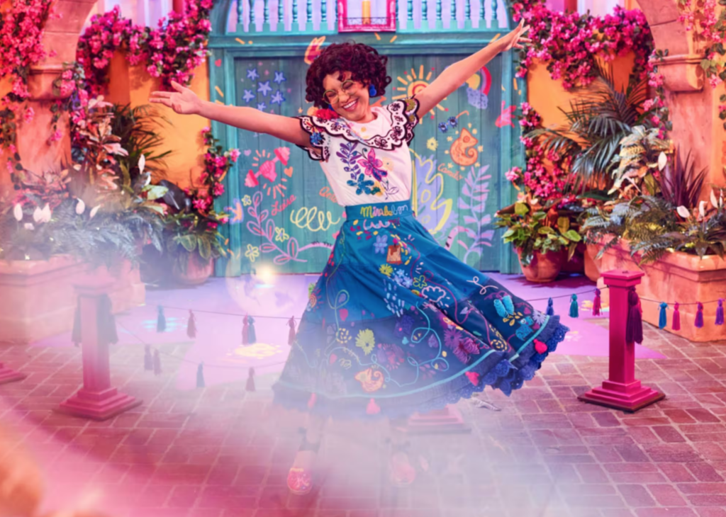 Mirabel from Encanto smiles big with her arms up in the air in an outdoor setting themed to her Madrigal house at Magic Kingdom