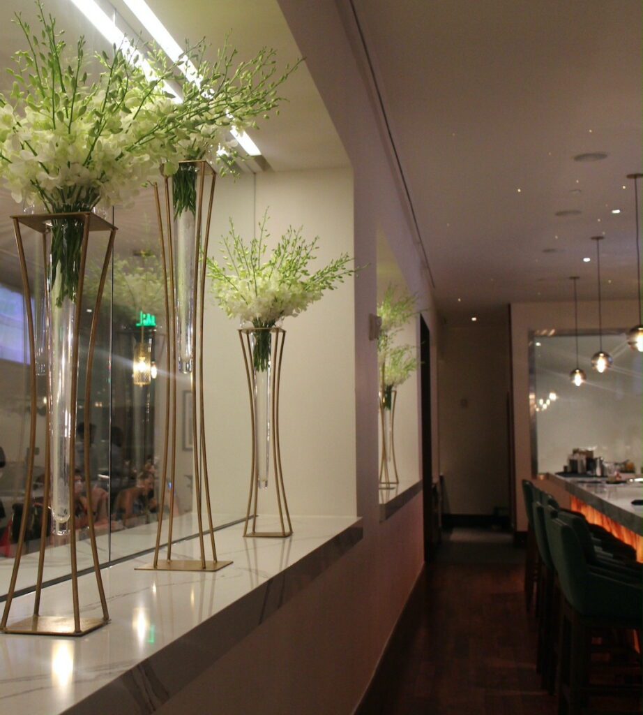 The Steak House 71 bar with large vases and light green floral behind the seats with a glass window separating the space from the restaurant portion of the venue