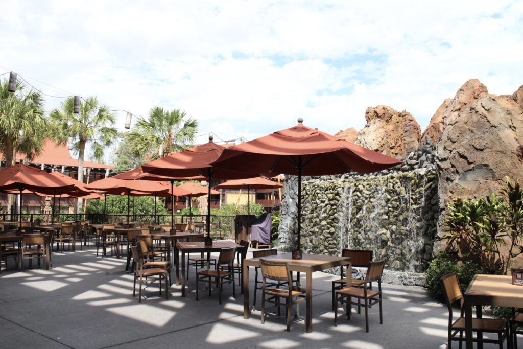 Outdoor tables and chairs with umbrellas in a tropical setting with a waterfall wall, palm trees and string lights