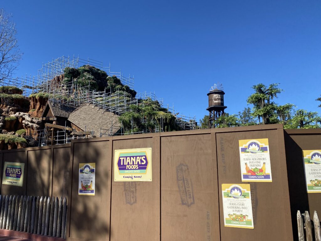 A brown construction wall with Tiana's foods business marketing on it. In the distance the former Splash Mountain attraction has been reshaped and covered in new foliage.