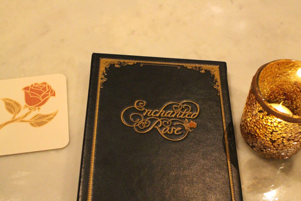A black Enchanted Rose menu cover with the bar and lounge name in gold script next to a rose coaster and a gold candle holder.