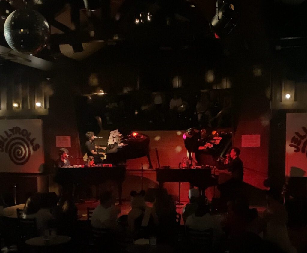 Two piano players play with instruments that face each other on a dark stage lit by a disco ball at Jellyrolls Disney location.