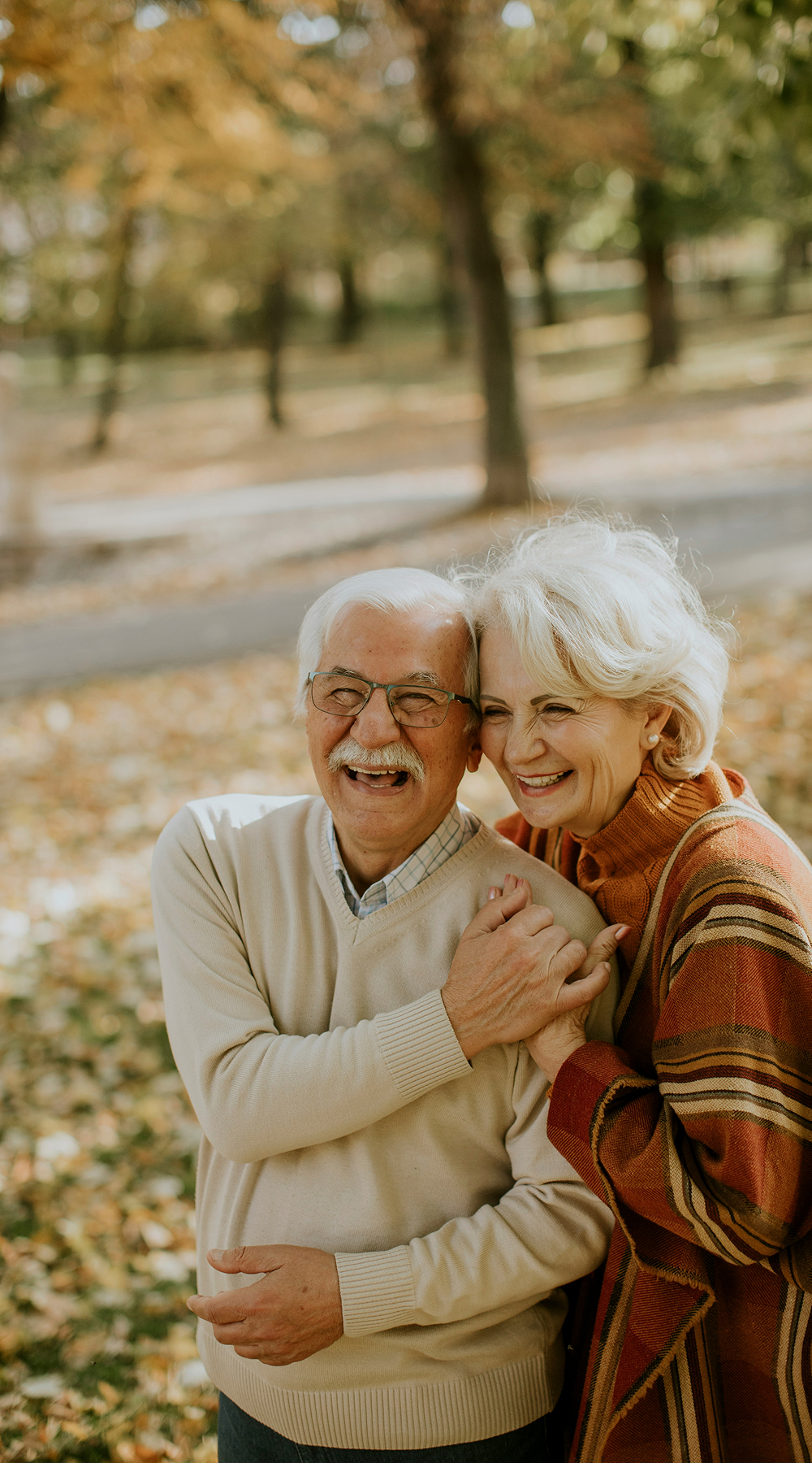 Smiling elderly couple embracing in autumn park with fallen leaves and trees in background.