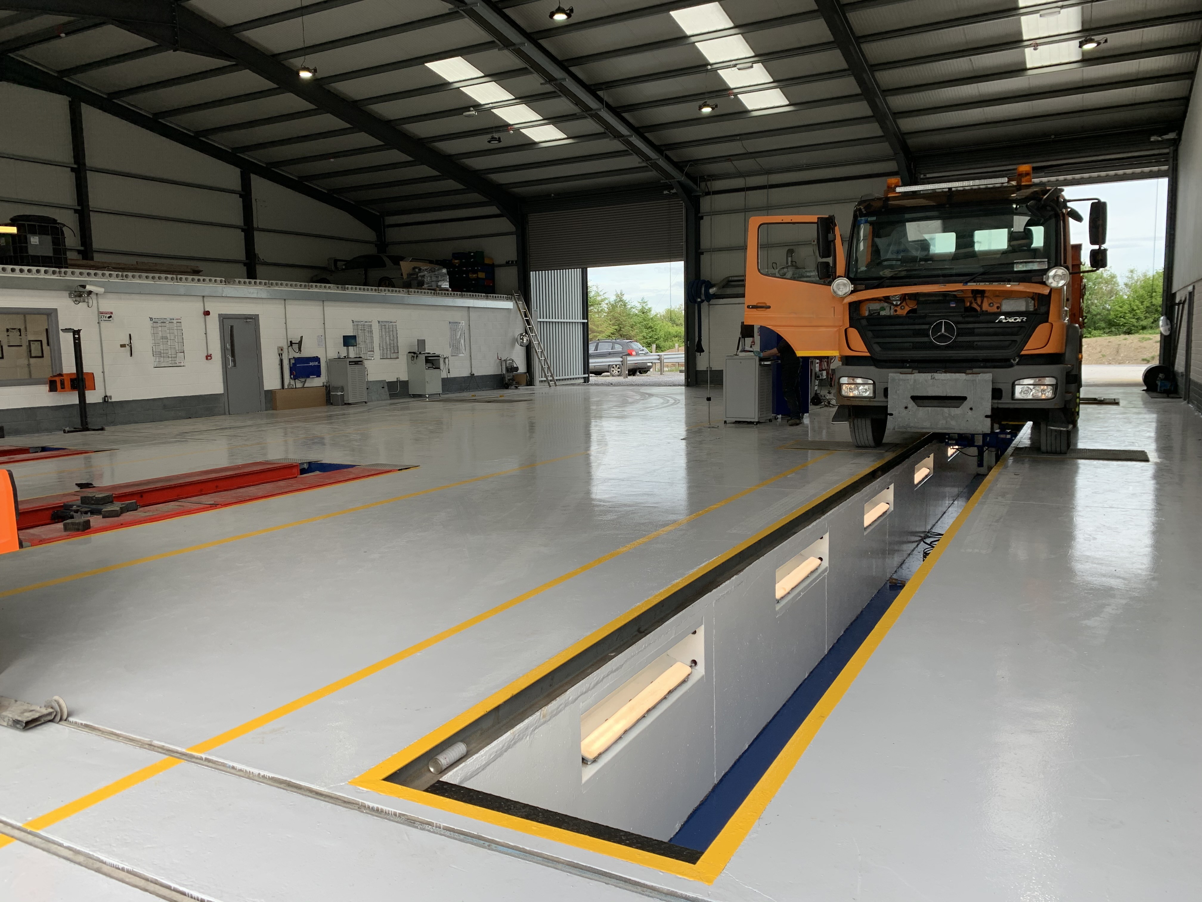 Orange Mercedes truck at a garage inspection pit inside a clean, spacious vehicle service bay.