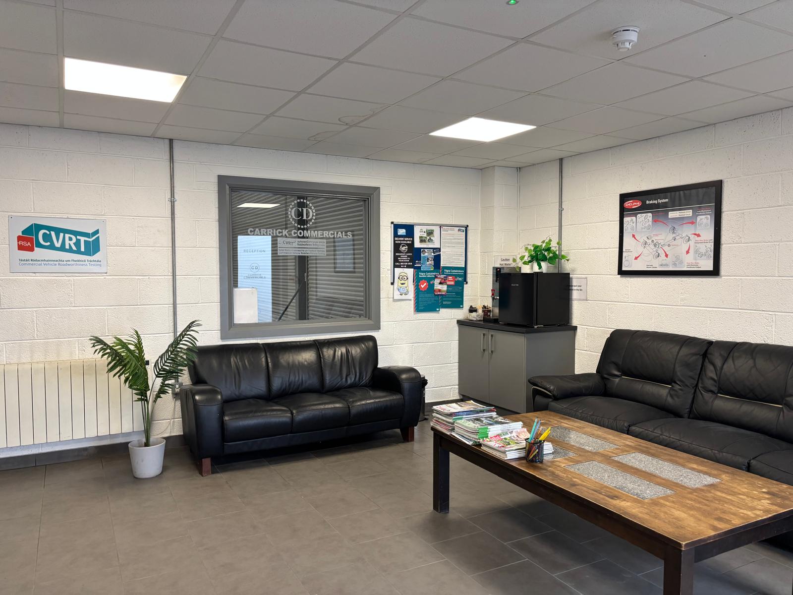 Waiting room with two black leather sofas, a wooden coffee table, potted plant, and a coffee machine cabinet.