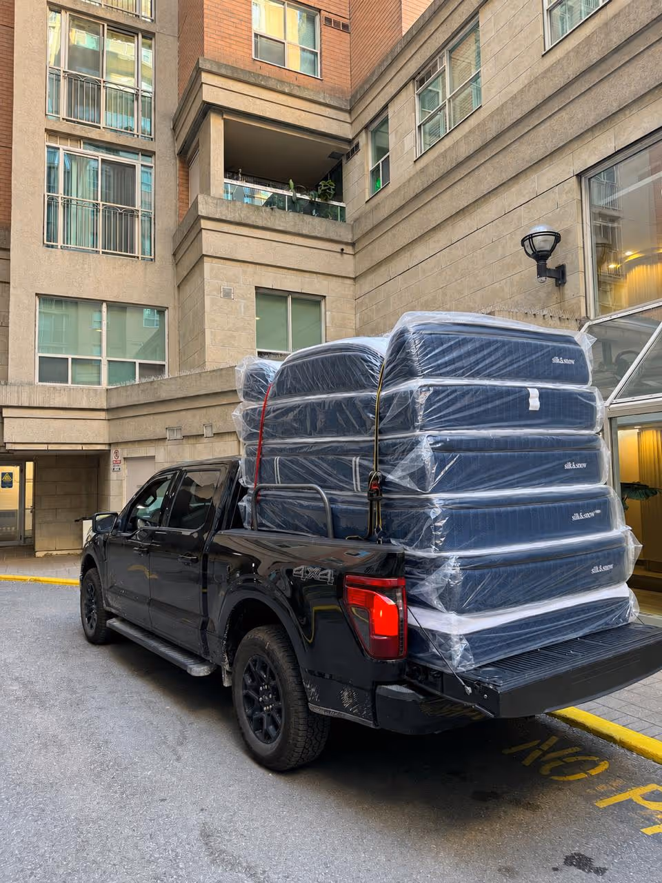Three people carrying a wrapped mattress outside a warehouse with trucks in the background.