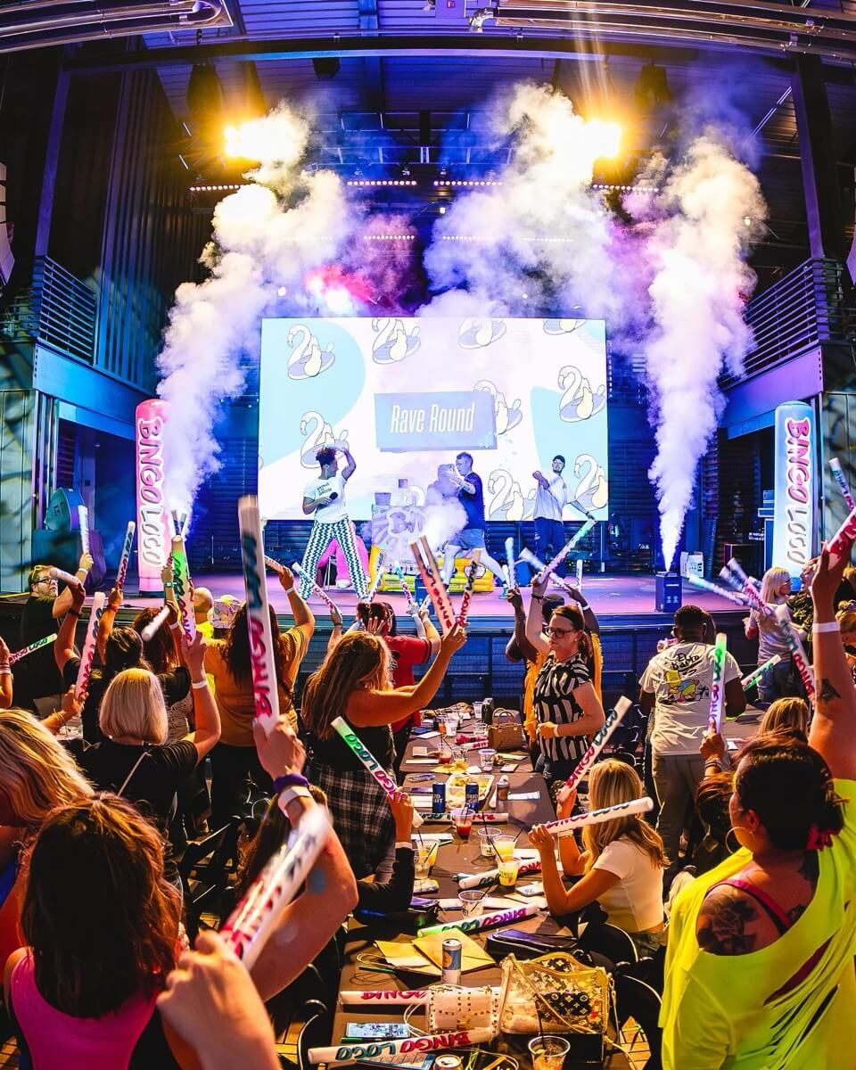 Crowd waving light sticks at a lively indoor event with stage, performers, and smoke effects.