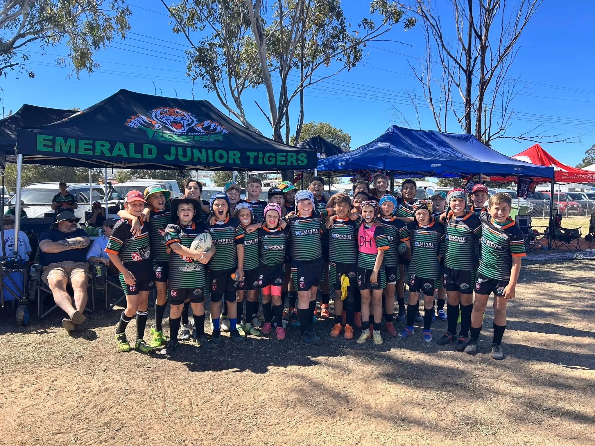 Group of youth rugby players in black and green Emerald Junior Tigers uniforms posing outdoors under tents.
