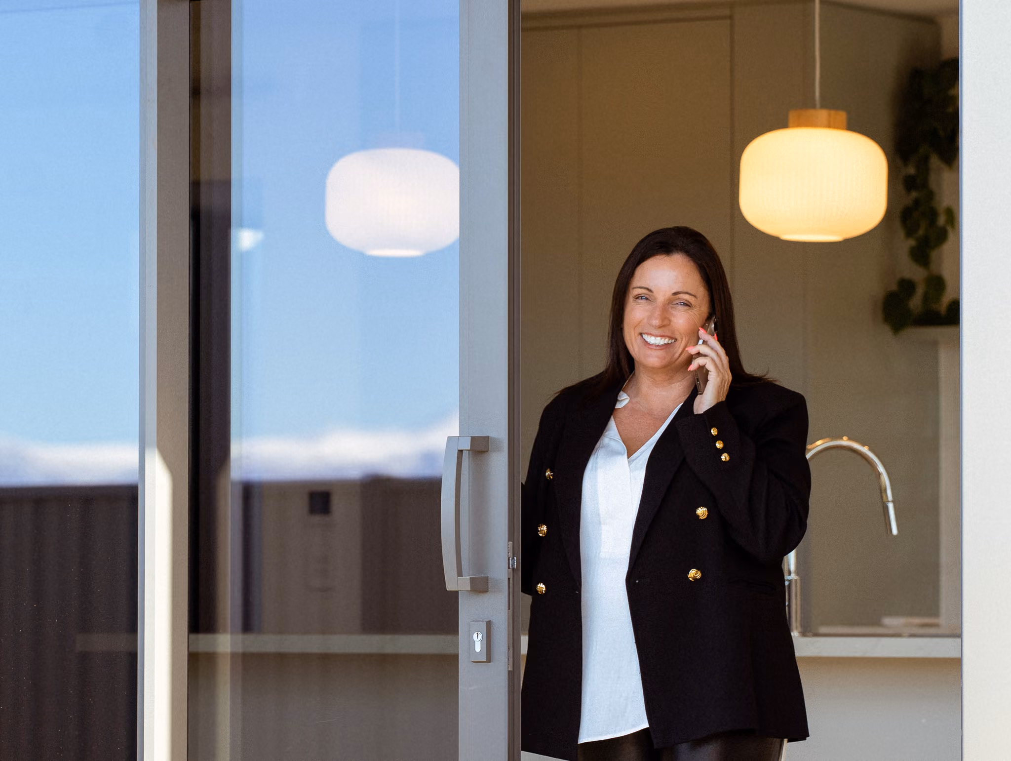 Coralie standing in an open sliding door, smiling at the camera while on the phone