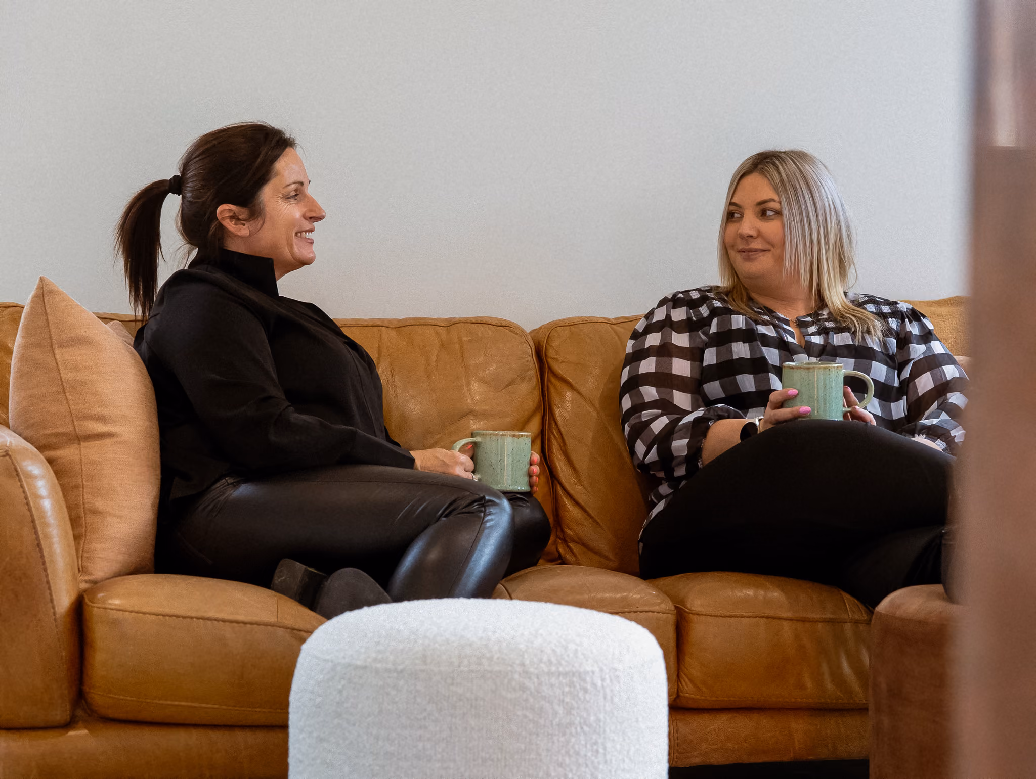 Coralie and Erica sitting on a couch with mugs while smiling at eachother