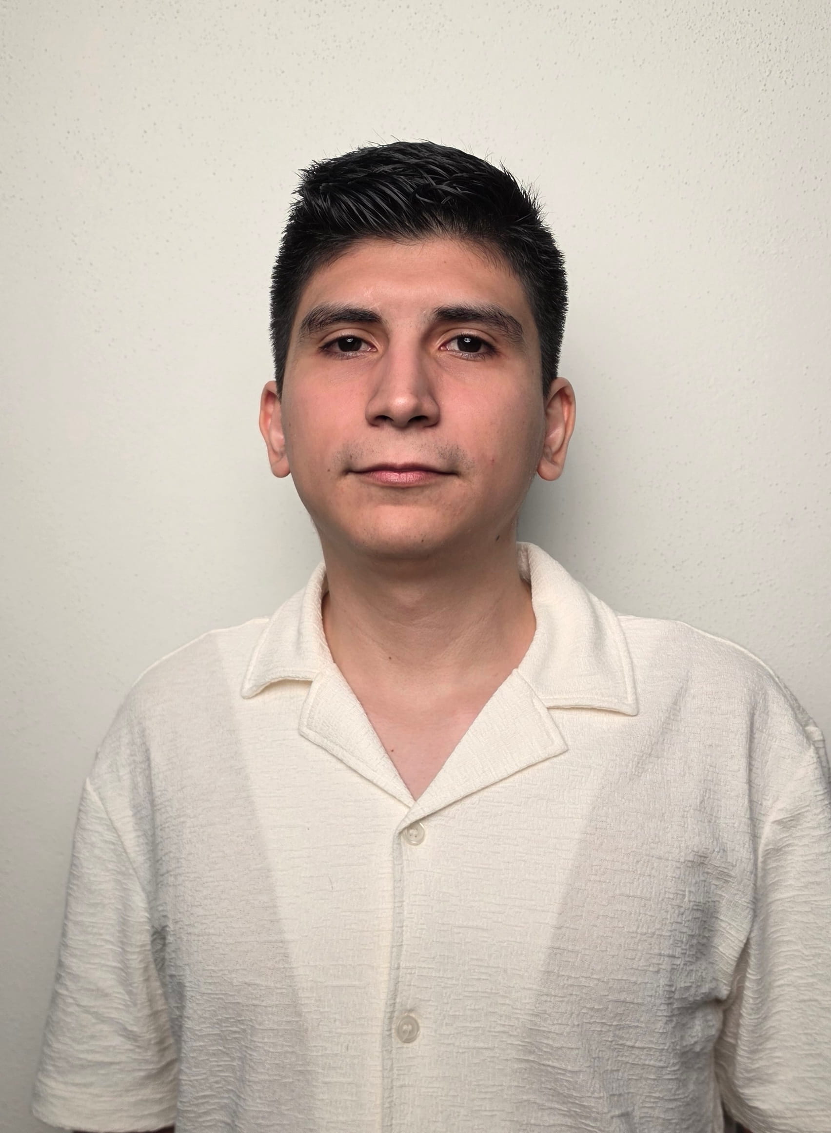 Hombre joven con cabello negro corto y camisa blanca mirando al frente sobre fondo claro.