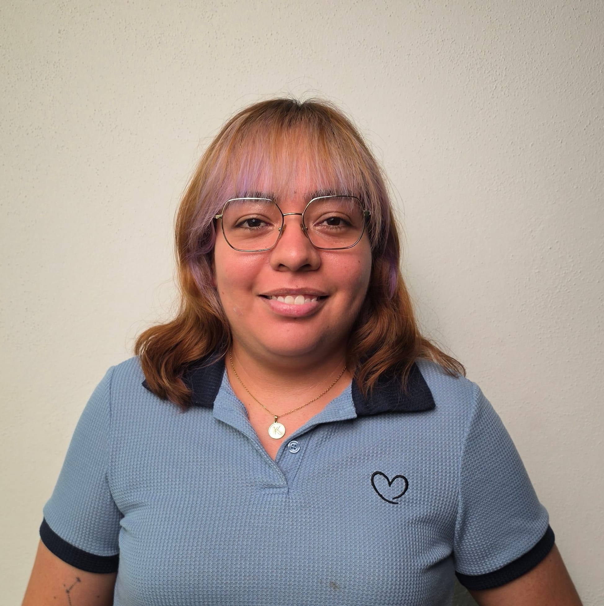 Mujer con gafas, cabello castaño con reflejos morados y camiseta azul con corazón negro.