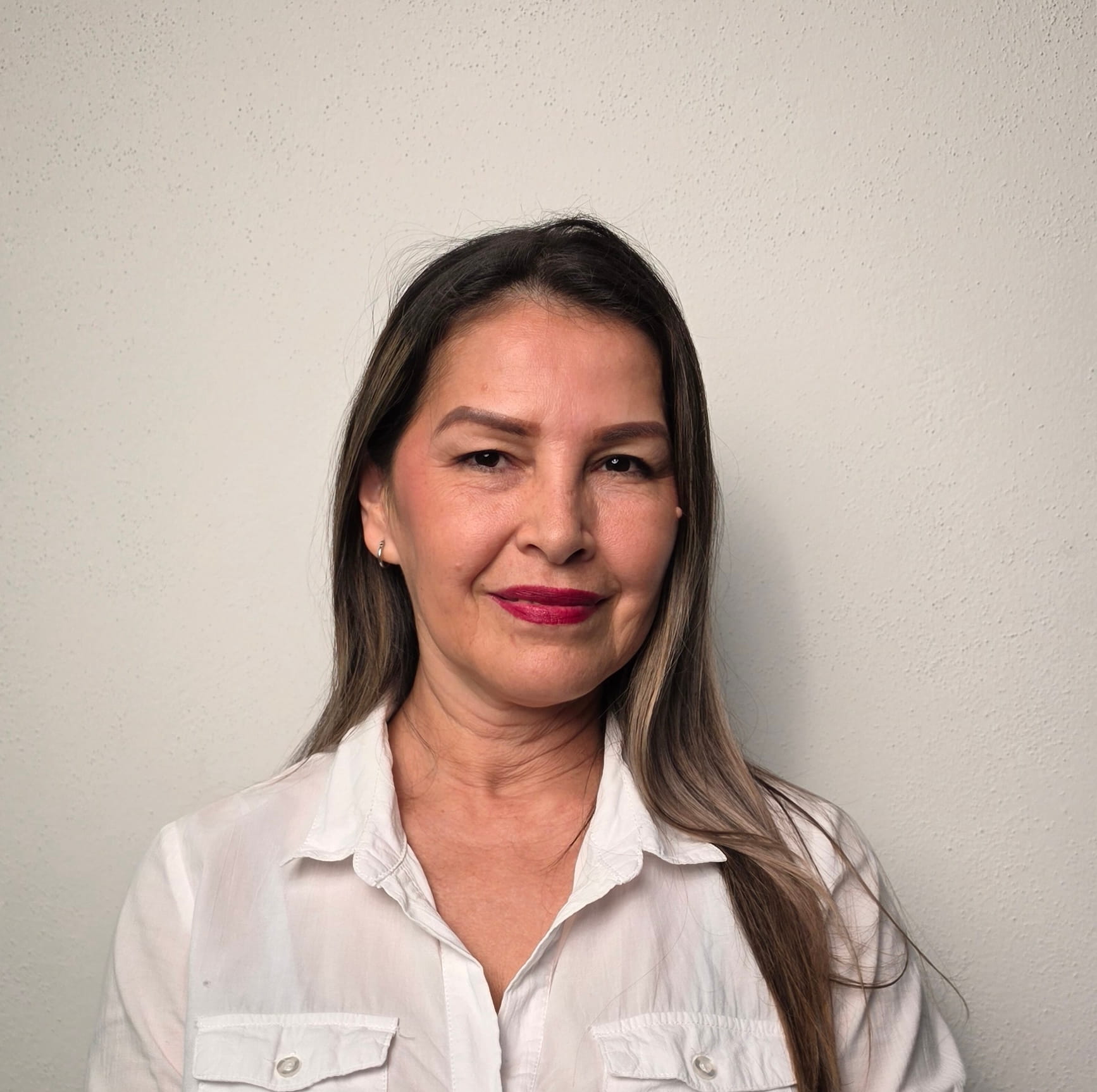 Mujer sonriente con camisa blanca y labios rojos frente a una pared beige.