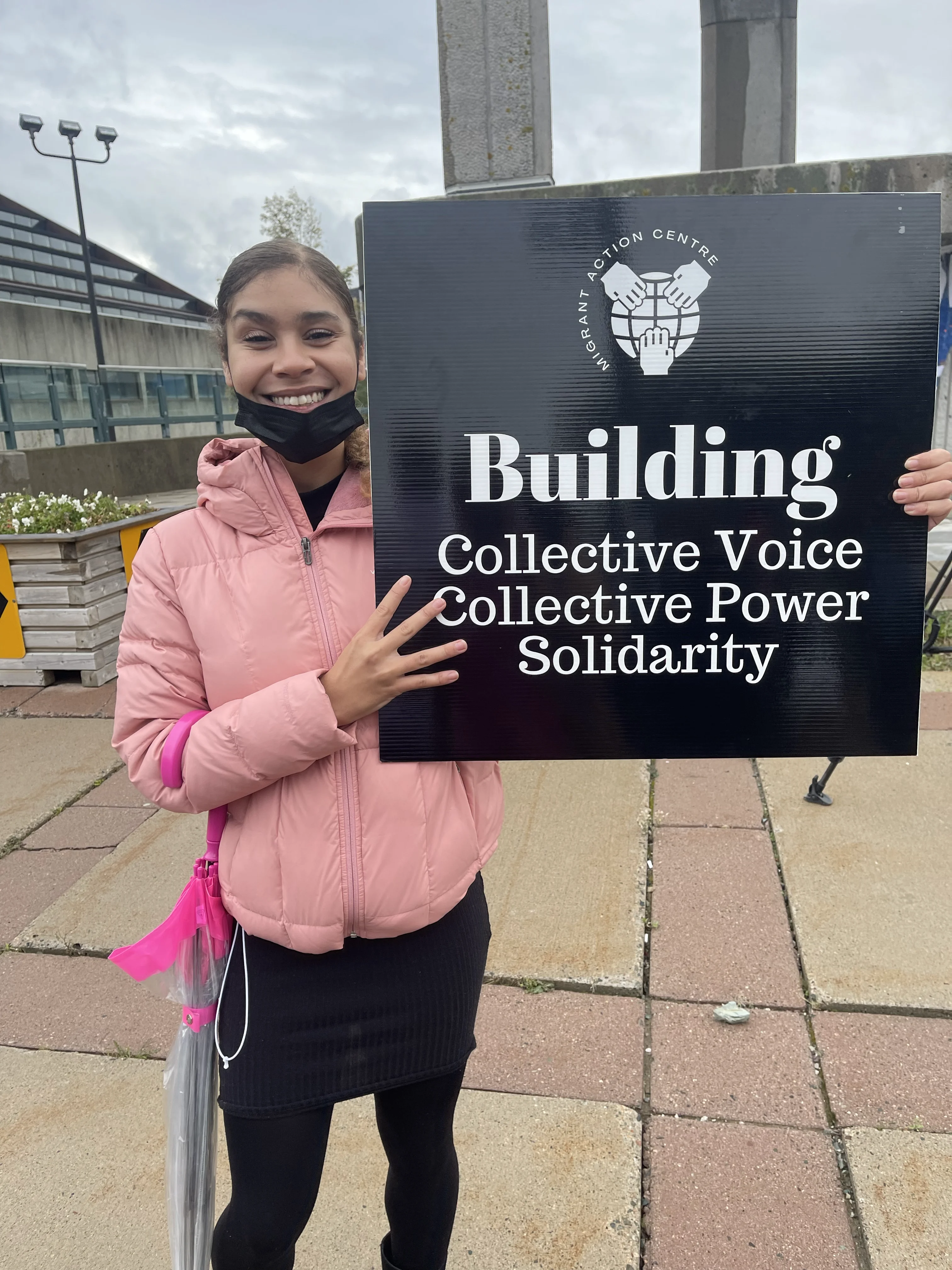 A person holding a sign that features a square black placard with a white Migrant Action Centre logo and text underneath that reads "Building Collective Voice Collective Power Solidarity"