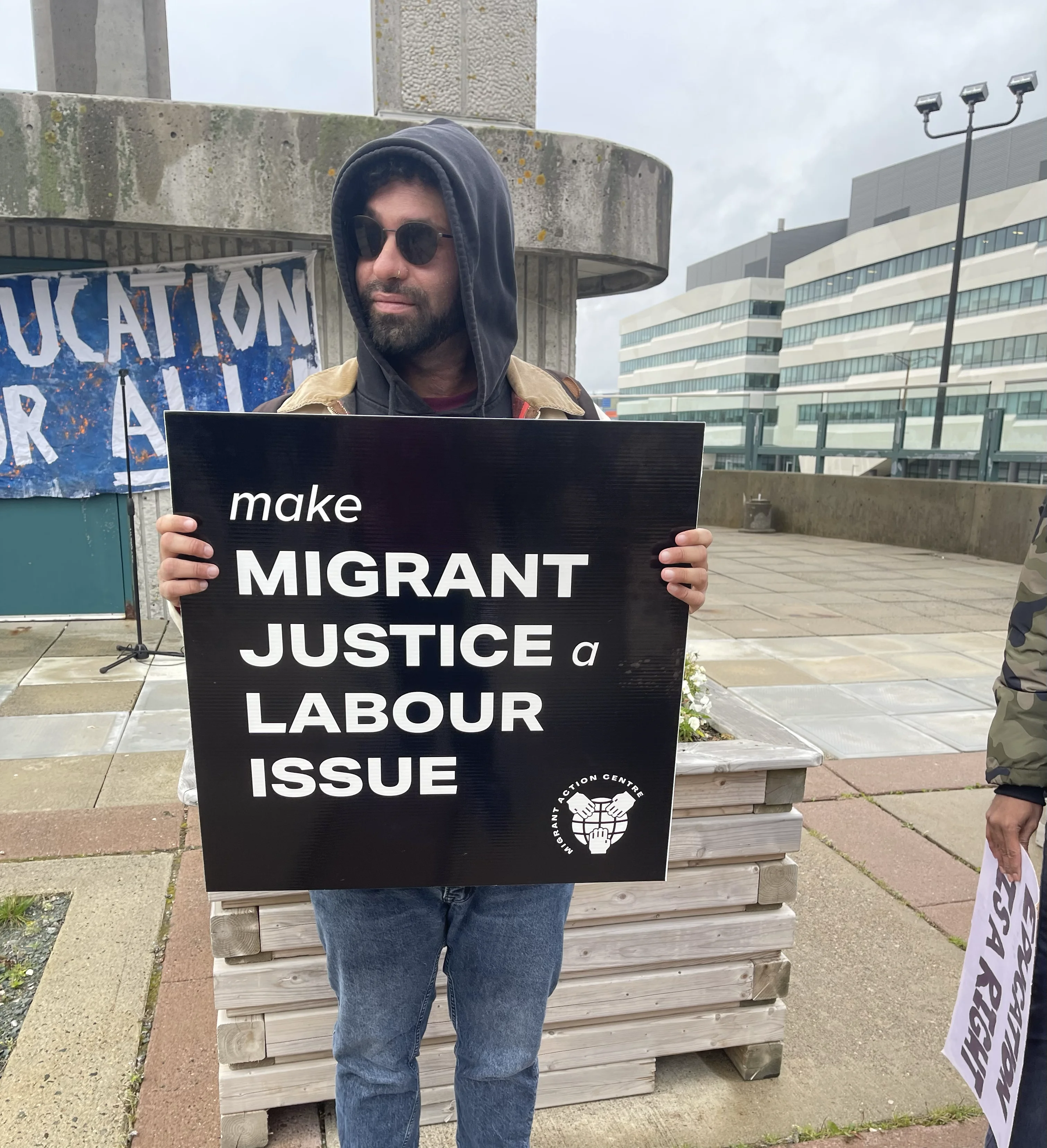 A person holding a square sign with a black background and white text that reads "make migrant justice a labour issue" and features a white Migrant Action Centre logo on the bottom right corner.