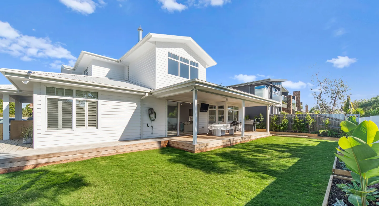 Modern two-story white house with large windows and a covered wooden patio overlooking a well-maintained green lawn and garden under a clear blue sky.