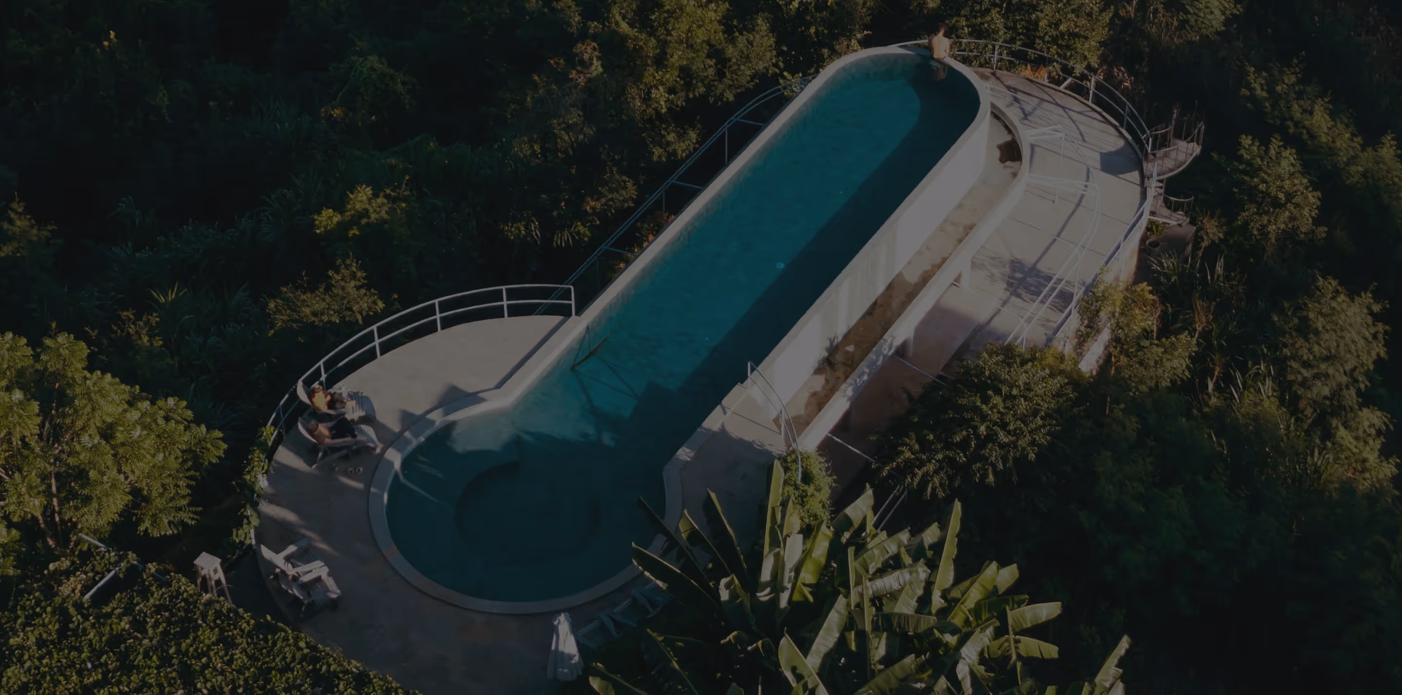 A long, narrow pool surrounded by dense green trees and two people sitting on chairs nearby.