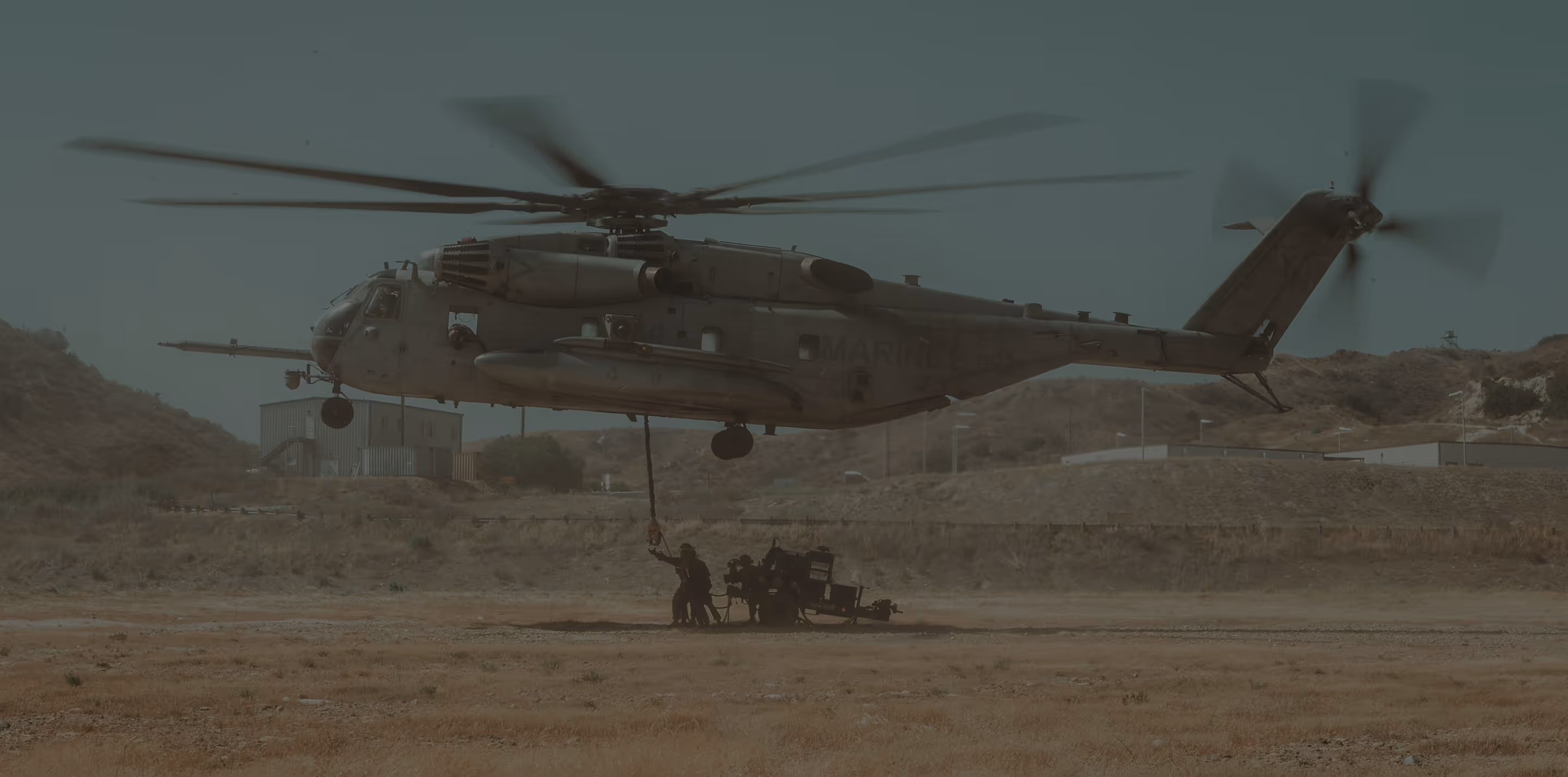 Military helicopter hovering close to ground with soldiers rappelling from a rope in a dry terrain area.