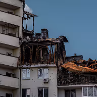 Partially burned apartment building with charred upper floors and intact lower floors under clear sky.