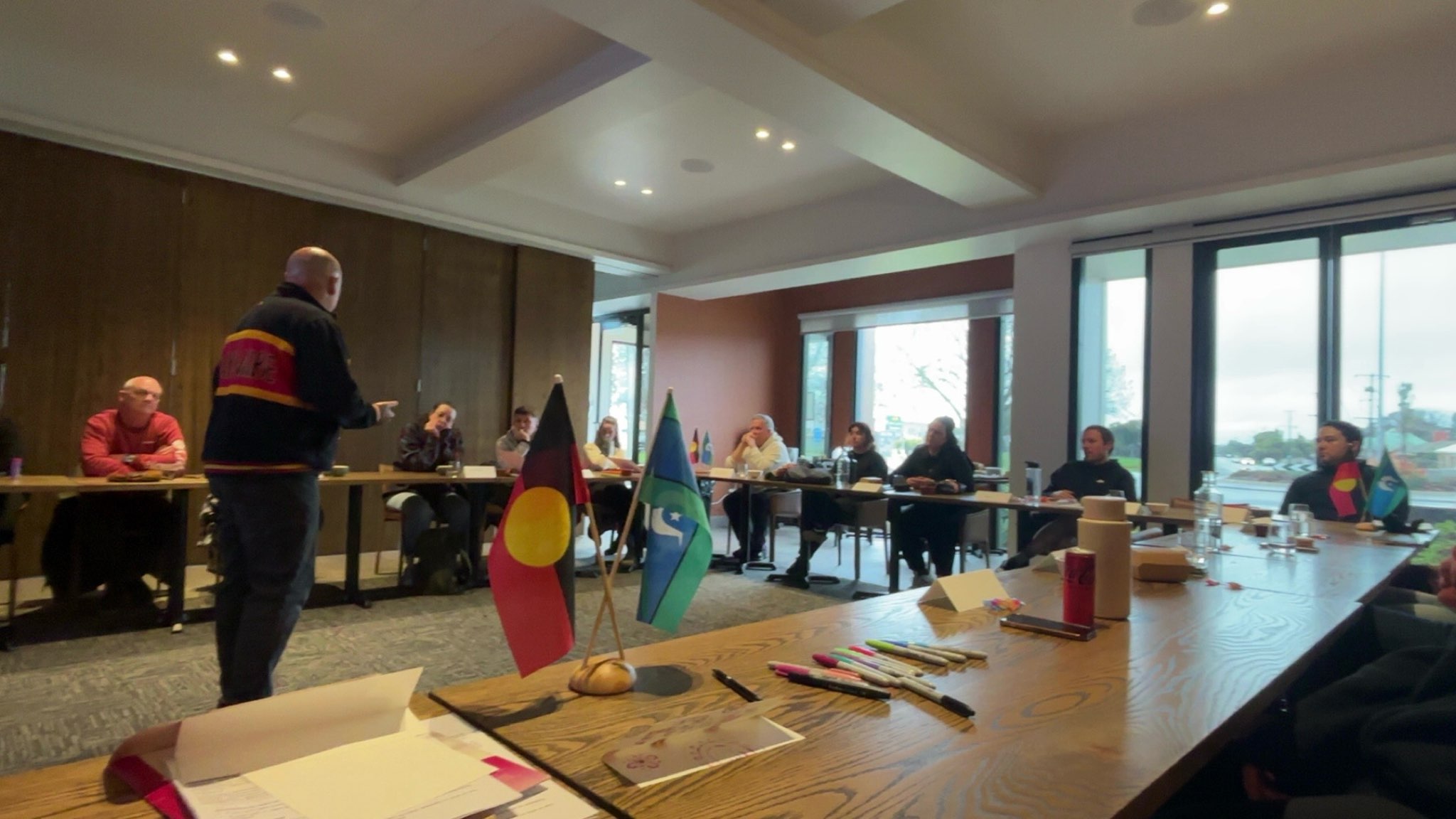 Shawn Andrews facilitating a Be BRAVE Feel SAFE session, the Aboriginal and Torres Strait Islander flags on the table, participants around the room