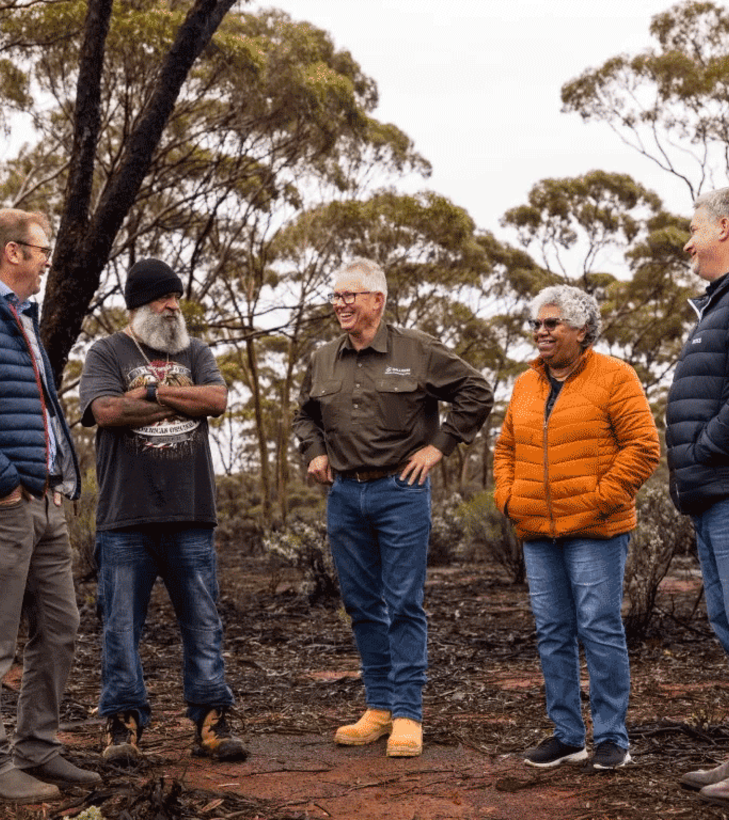 Five people standing outdoors in wooded area, smiling and talking, wearing casual and warm clothing.