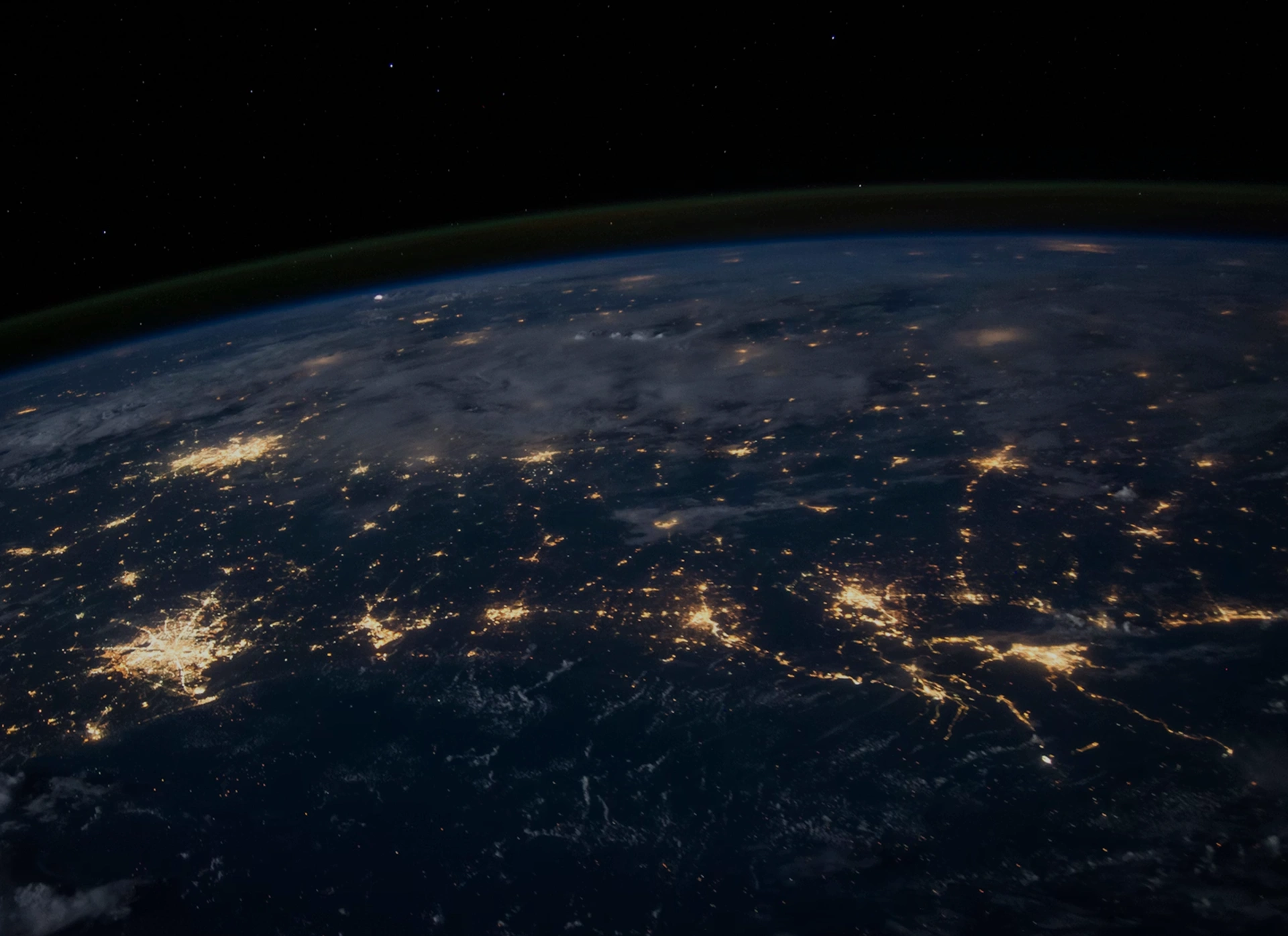 View of Earth at night from space showing city lights and cloud cover beneath a starry sky.
