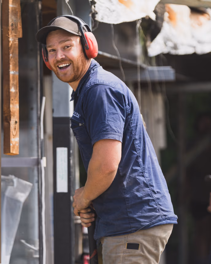 Smiling construction worker wearing red ear protection and a cap, holding a tool while working.