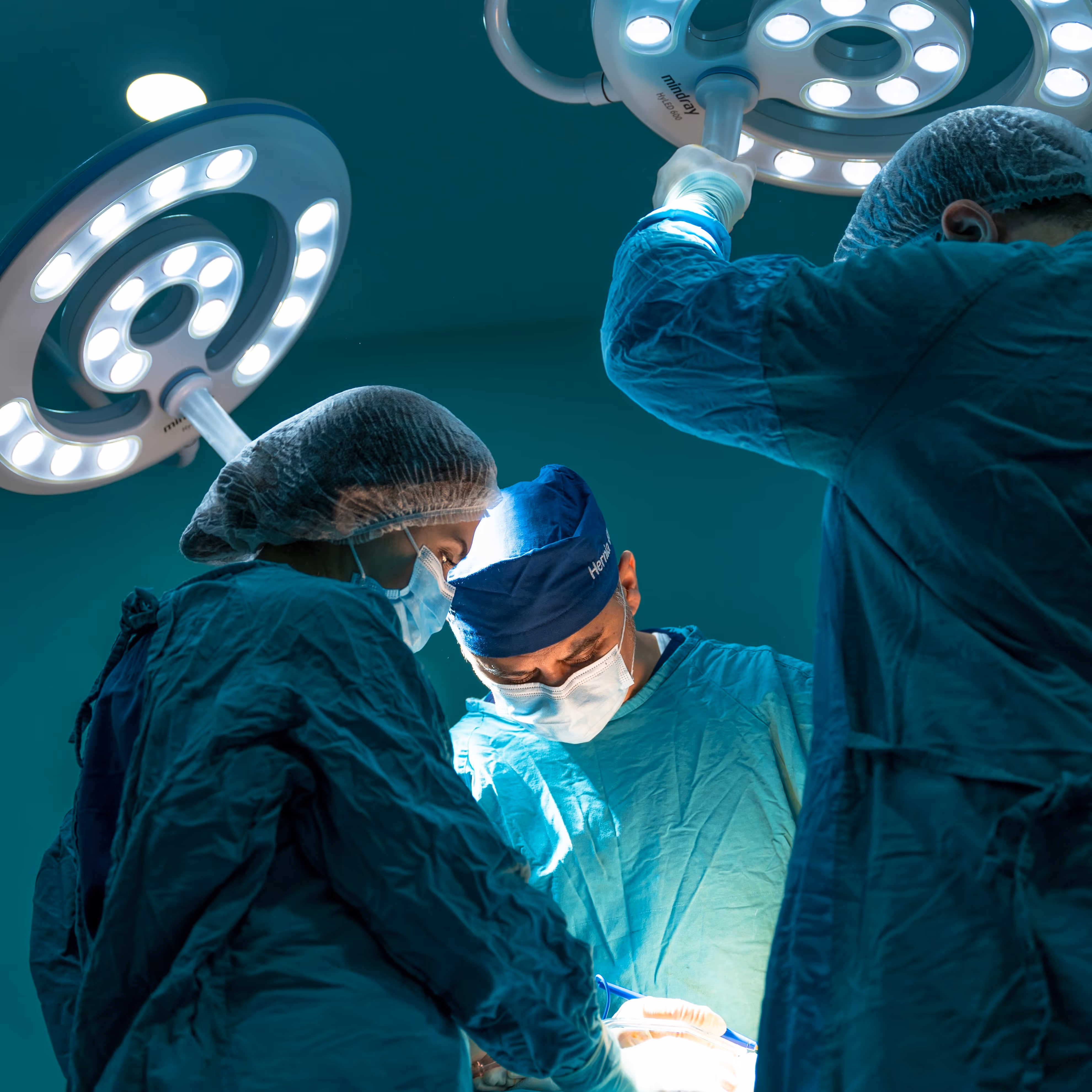 Trois chirurgiens en blouse opérant sous lumière chirurgicale dans une salle d'opération.