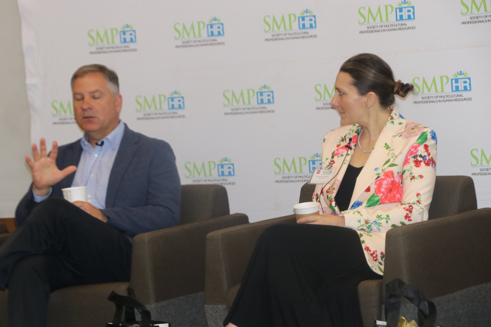 Two panelists in seated discussion at SMPHR event with branded backdrop