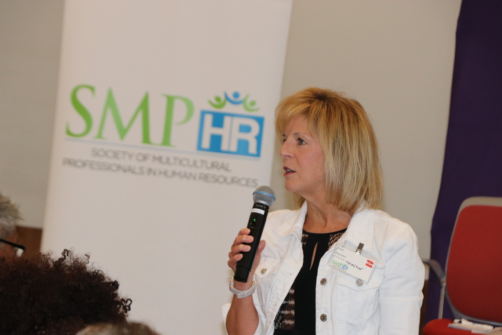 Woman in white blazer speaking at microphone with SMPHR logo visible