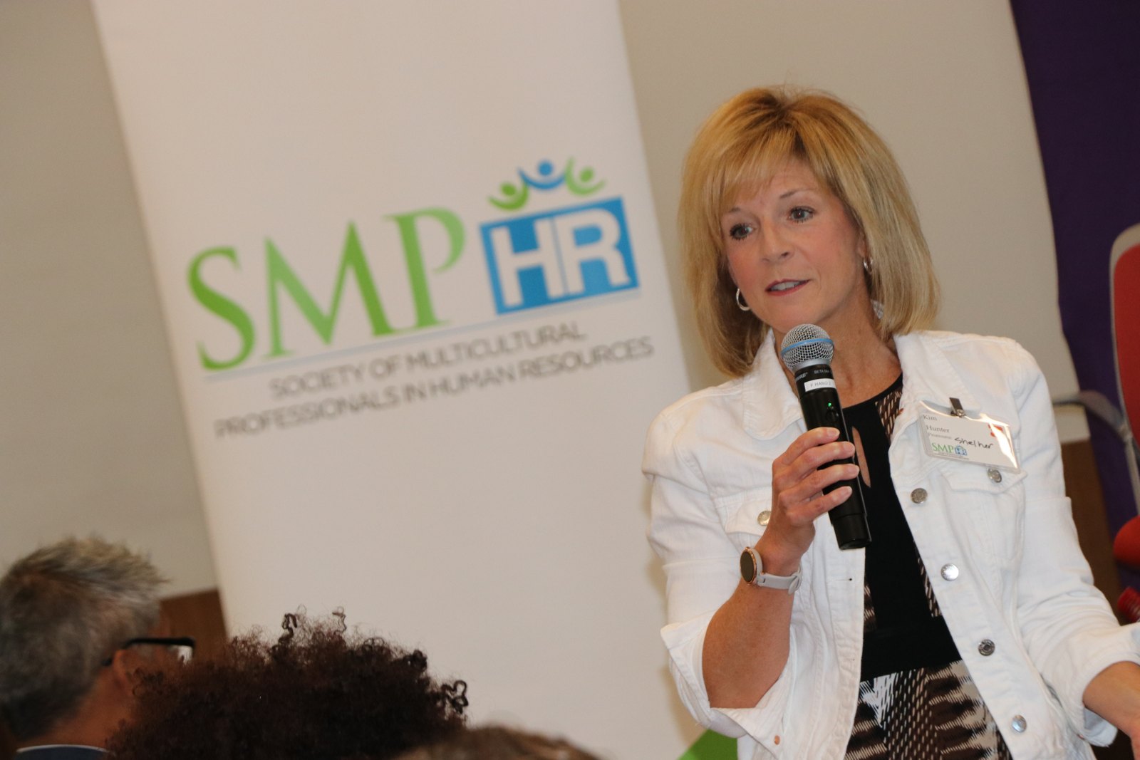 Woman in leopard print jacket speaking at microphone with audience members visible
